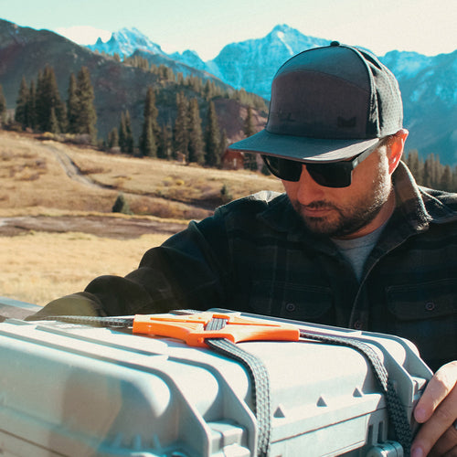 Man in outdoor setting with mountains, holding a large gray cooler with orange handles securing with black and orange Power Strap.