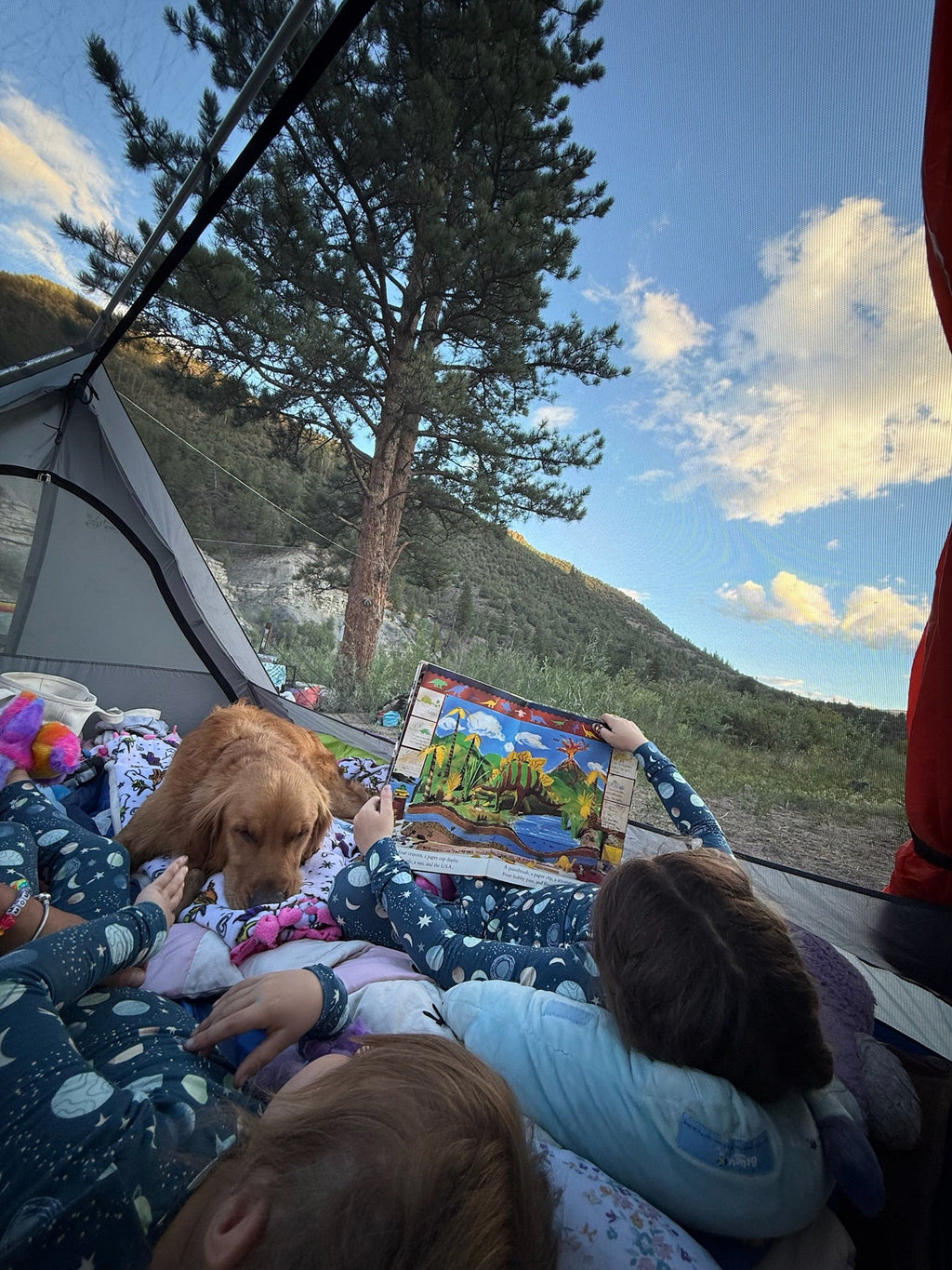 A cozy camping scene inside a tent with children and a golden retriever dog. One child is holding up a colorful landscape picture card, surrounded by pajamas, with a mountain and pine tree visible outside under a blue sky.