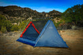 A blue and red tent pitched outdoors in a dry mountain landscape under a blue sky, with trees and rocky hills in the background.